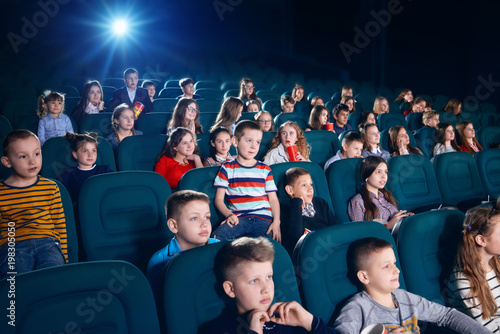 Sideview of children watching movie in the cinema hall. Kids are very emotional, exited and satisfied. Some children eating popcorn or drinking fizzy drinks.