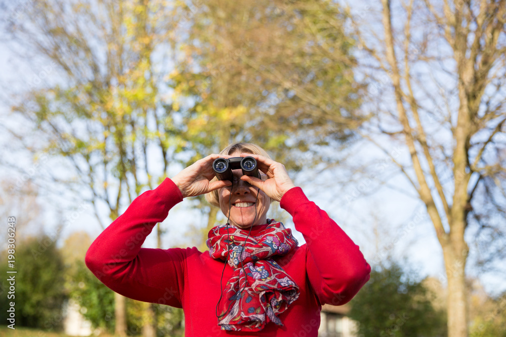 Smiling mature woman looking through binoculars in forest Smiling mature woman looking through binoculars in forest