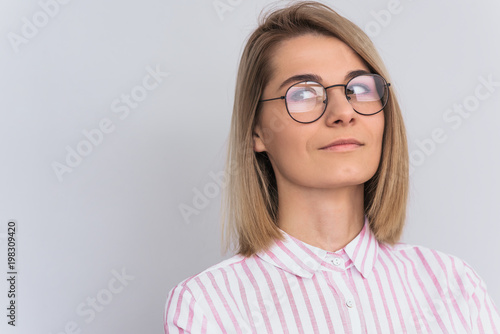 Portrait of positive blonde female with attractive look wearing pink shirt and round trendy glasses, posing against white wall in studio. Happy woman showing positive emotions looks aside.