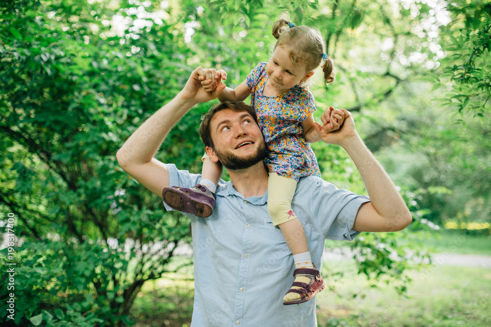 Fototapeta premium Father and daughter playing piggyback at outdoor garden park. Cute little blonde girl and man with a beard having fun