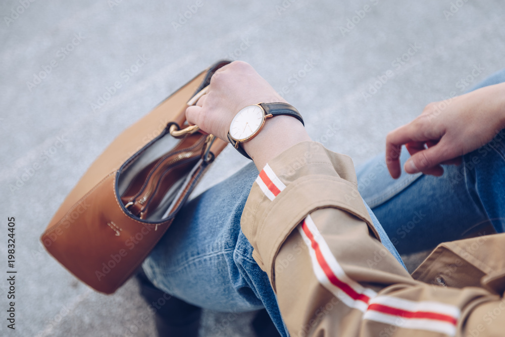 outfit details, chic woman wearing a stylish beige trench coat and a black and white wrist watch. detail shot of a fashion bloggers outfit, accessorized with a metallic handle brown leather tote bag.