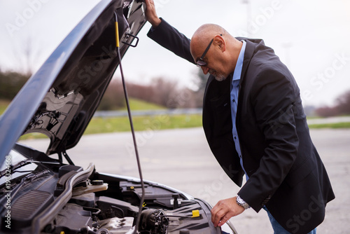 Mature professional elegant worried businessman in the suit is looking under the car hood trying to figure out the problem.