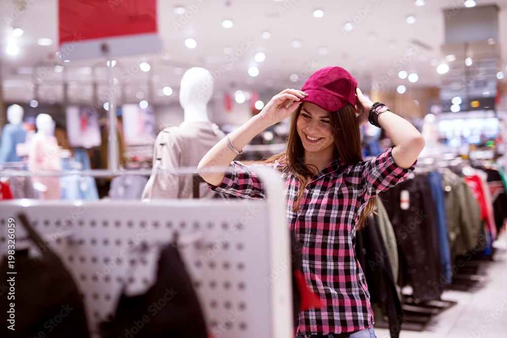 Young gorgeous happy girl is browsing and trying out blue hat while standing in a store. Young gorgeous happy girl is browsing and trying out blue hat while standing in a store.