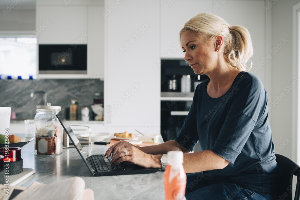 Side view of mature blond woman using laptop at kitchen island Side view of mature blond woman using laptop at kitchen island