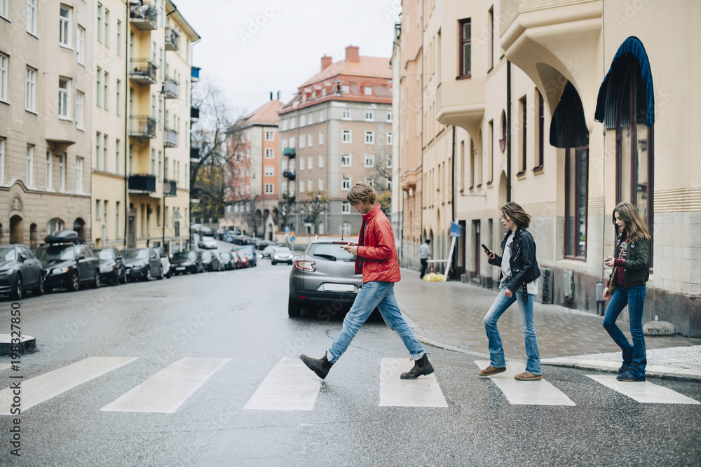 Full length of friends using mobile phone while crossing street in city Full length of friends using mobile phone while crossing street in city