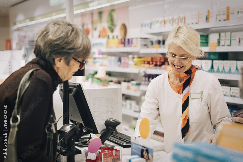 Smiling female owner standing with senior customer at checkout in pharmacy store Smiling female owner standing with senior customer at checkout in pharmacy store