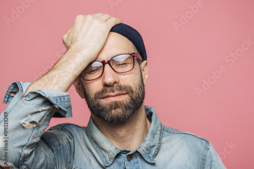 Portrait of depressed middle aged unshaven male closes eyes and keeps hand on forehead, has unhappy expression, isolated over pink background. Fatigue hipster guy has headache after hard work