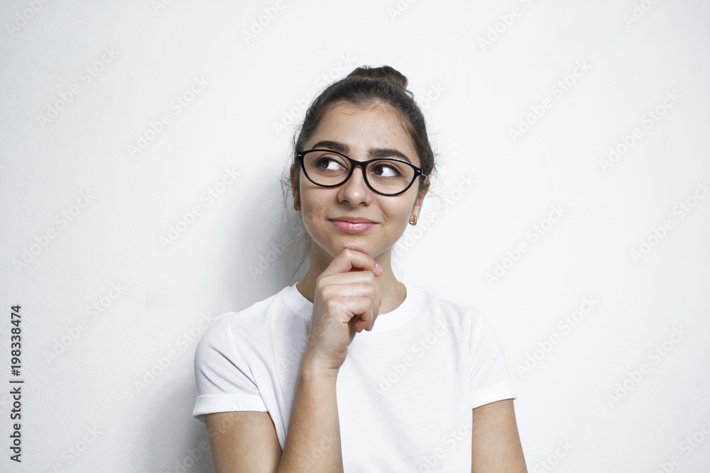 A thoughtful, positive Indian girl in glasses thinks about the idea. A student on a white background dreams about something. - obrazy, fototapety, plakaty 