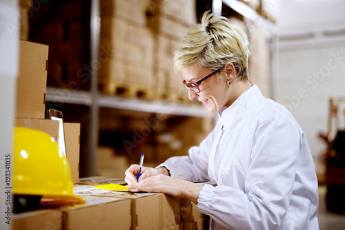 Young beautiful happy female worker is filling a paperwork on a stack of cardboard boxes while smiling in factory storage room.