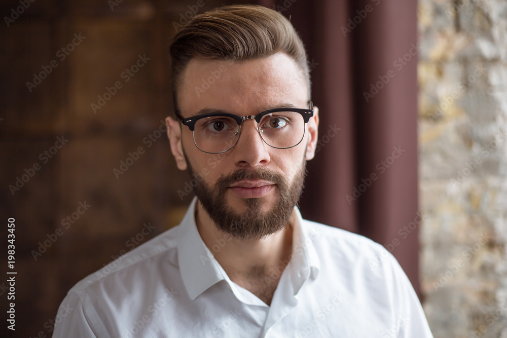 Young smiling handsome bearded businessman in a white shirt and glasses stands at the center of the office and looks at the camera. Young smiling handsome bearded businessman in a white shirt and glasses stands at the center of the office and looks at the camera.