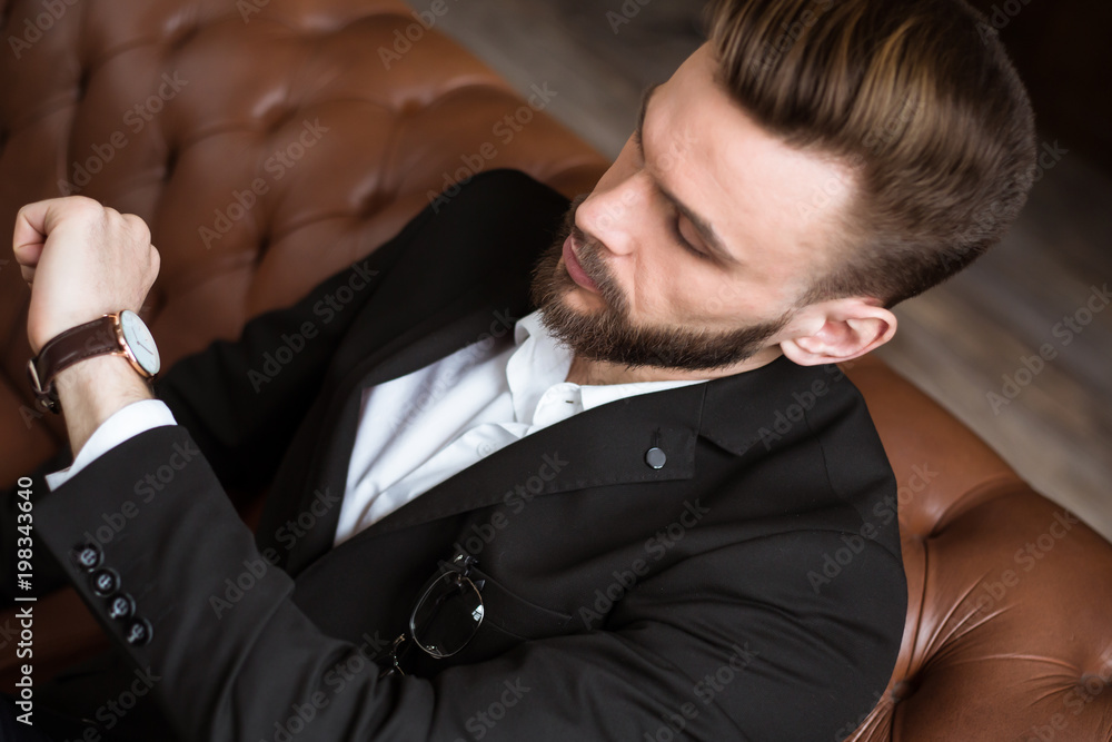 Time for work. Stylish handsome confident bearded businessman in smart casual wear sitting in office on sofa and looking at wristwatch. Time for work. Stylish handsome confident bearded businessman in smart casual wear sitting in office on sofa and looking at wristwatch.