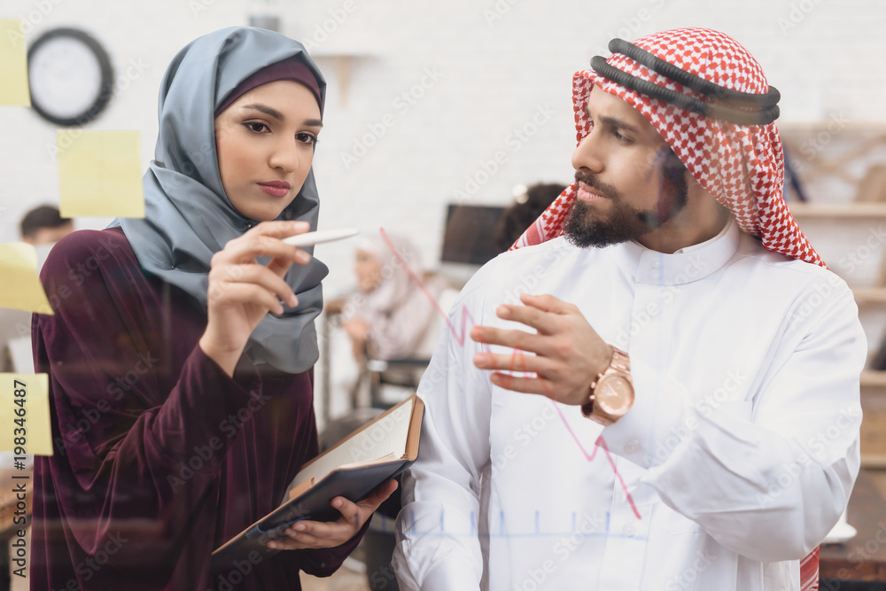 Arab man and woman working in offce. Coworkers are taking notes on glass board. Arab man and woman working in offce. Coworkers are taking notes on glass board.