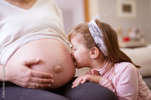 Close up view of lovely happy little toddler girl kissing shirtless pregnant tummy of her mother while resting at home.