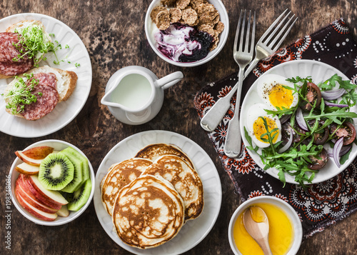 Greek yogurt with whole grain cereals and berry sauce, pancakes, arugula, cherry tomatoes, boiled eggs salad, kiwi, apples fruit, salami and cream cheese sandwiches on a wooden background, top view