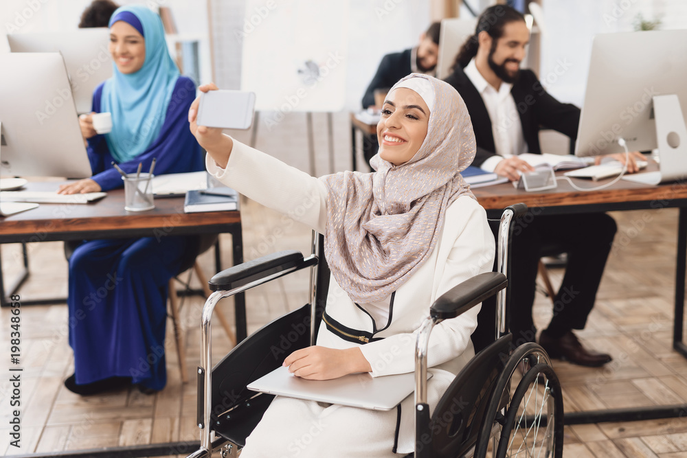 Disabled arab woman in wheelchair working in office. Woman is taking selfie.