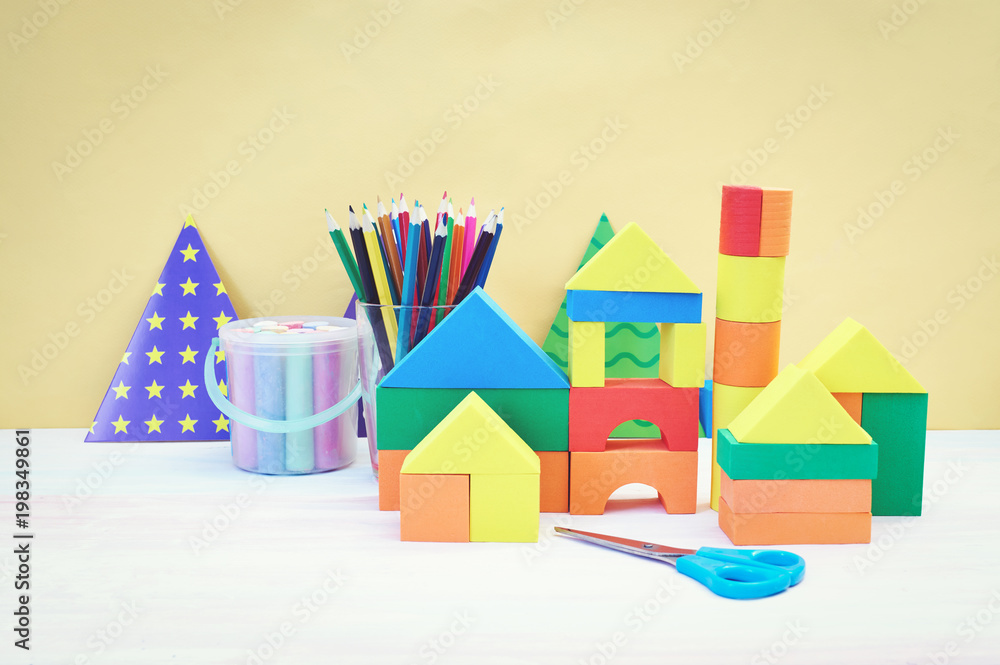 Multicolored festive triangular flags, wooden toy house and glass cup with pencils yellow background