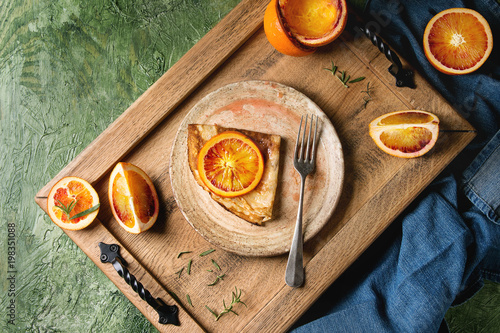 Homemade crepes pancakes served in ceramic plate with bloody oranges and rosemary syrup with sliced sicilian red oranges on wooden tray over green texture background. Top view, space