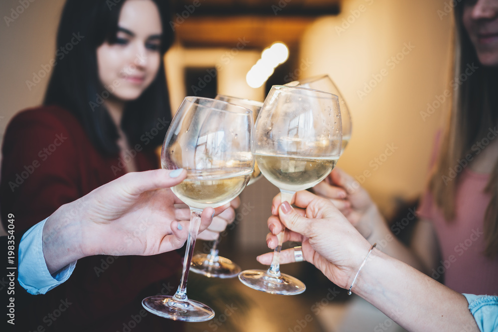 Group of happy friends making cheers with glasses of white wine, girlfriends celebrating and toasting at cozy home atmosphere, togetherness and traditional concept