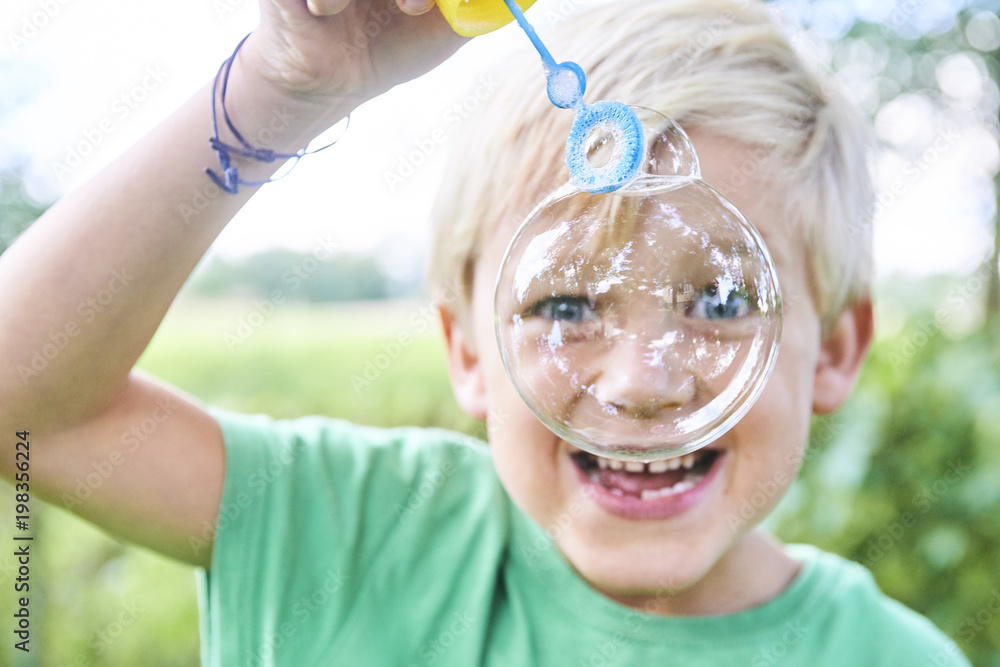 Young boy blowing bubbles with greenery background. Selective focus.
Young boy blowing bubbles with greenery background. Selective focus.