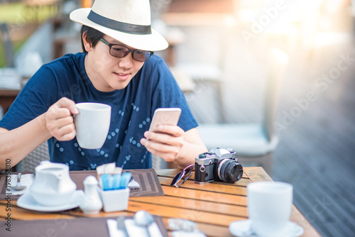 Young Asian man holding a cup of coffee using smart phone during having breakfast in resort  coffee time and summer holiday vacation concepts