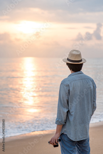 Young Asian man traveler and photographer with jean shirt and hat holding film camera and looking beautiful sunset at tropical beach island. Summer holiday or vacation travel concepts © zephyr_p