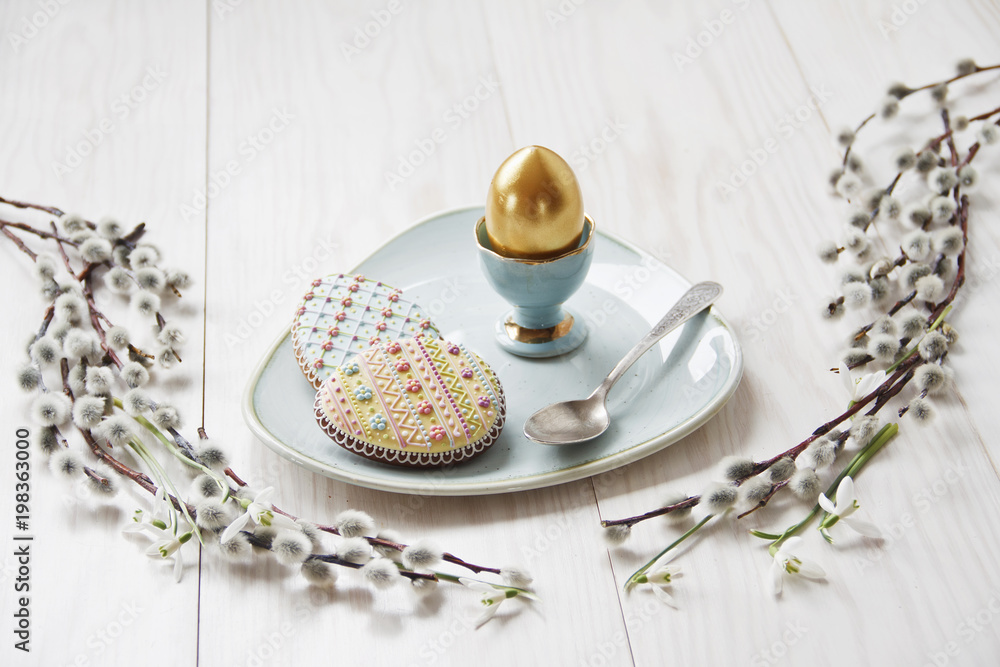 Close-up photo of Easter golden egg on plate with cookies and metal spoon on wooden background with willows branches Close-up photo of Easter golden egg on plate with cookies and metal spoon on wooden background with willows branches