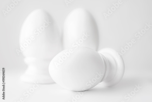 Interior home or church decor of three white ceramic eggs on stands or in egg-cups on white blurred background. Easter religious Christian symbol. Monochrome image.