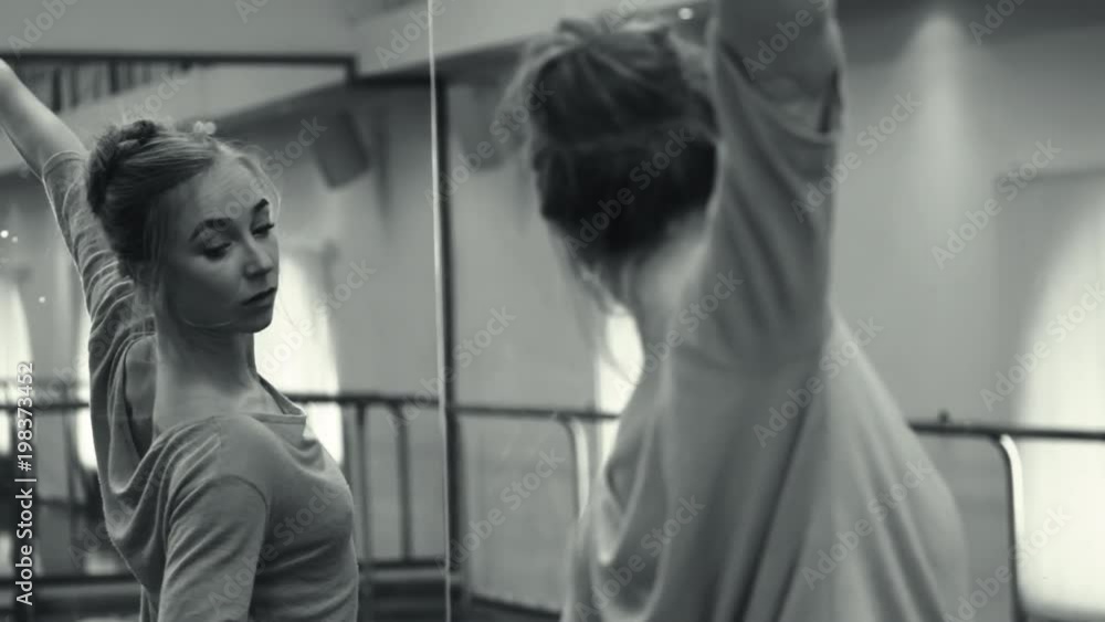Ballerina in beige sweatsuit and pointe stretches on barre in ballet gym. Woman standing near bar and mirror, preparing for performance or kneading before the lesson. Black white Ballerina in beige sweatsuit and pointe stretches on barre in ballet gym. Woman standing near bar and mirror, preparing for performance or kneading before the lesson. Black white