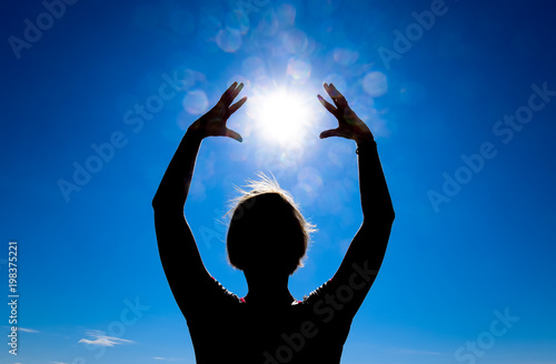 Silhouette of a girl against the background of the sun and blue sky. Hands are raised up to the sun.