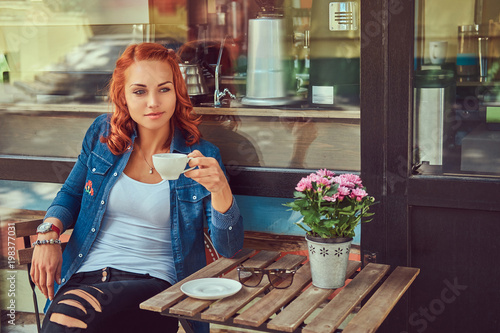 A beauty redhead girl drinks coffee  sitting near the coffee shop.