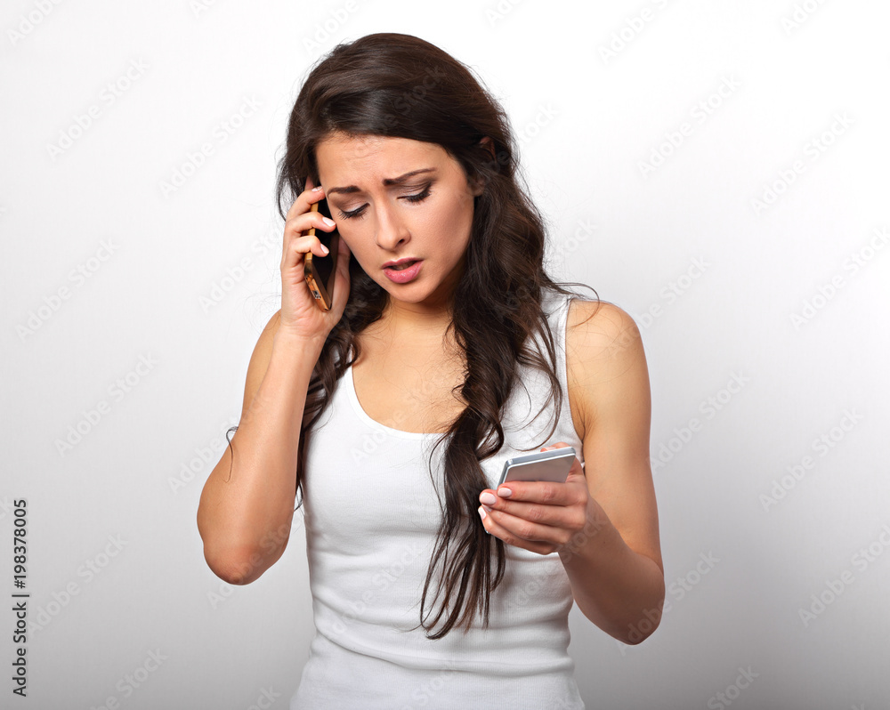 Worried unhappy young woman talking on one mobile phone and typing sms looking on two mobile phone on white background Worried unhappy young woman talking on one mobile phone and typing sms looking on two mobile phone on white background