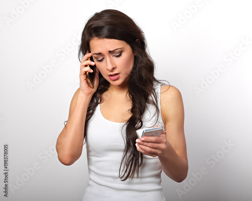 Worried unhappy young woman talking on one mobile phone and typing sms looking on two mobile phone on white background