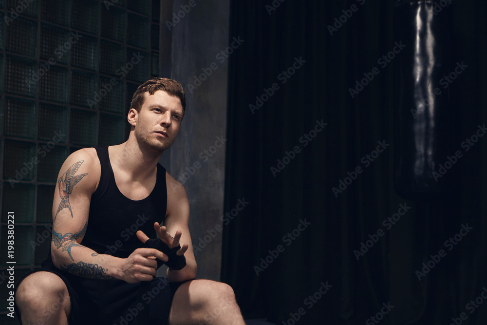 Indoor shot of thoughtful attractive young European man wearing sleeveless shirt and shorts wrapping boxing bandages, sitting against black curtain background with copyspace for your information Indoor shot of thoughtful attractive young European man wearing sleeveless shirt and shorts wrapping boxing bandages, sitting against black curtain background with copyspace for your information