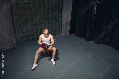 Top view of fashionable serious young muscular businessman wearing white sleeveless shirt, sneakers and red trousers taping hands with bandages before boxing training after working day at office