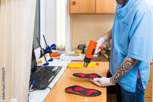 closeup Hands of young man with tattoo in workshop dressed in blue uniform make individual orthopedic insoles. The instrument uses hairdryer to heat and deform. Theme of small business and medicine.