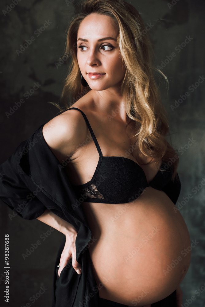 Fototapeta premium Pregnant woman. Portrait of an attractive pregnant woman with cute big belly in black lingerie who stands on the background of a dark wall