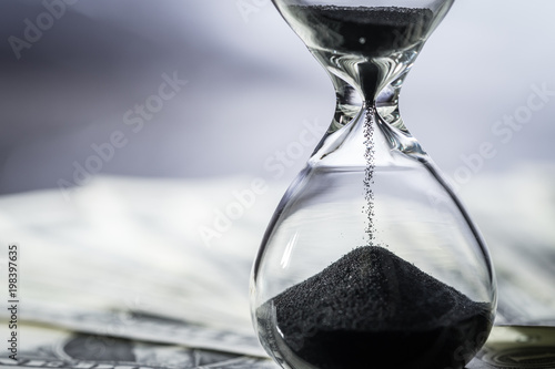 Closed up of sand falling in sandglass or hourglass on US Dollar bills as time running, long term investment or financial deadline concept