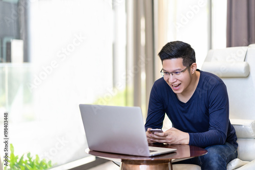 Happy excited asian man celebrate with smartphone and computer laptop, success or happy action. Freelancer or Entrepreneur using technology and start up modern office with copy space