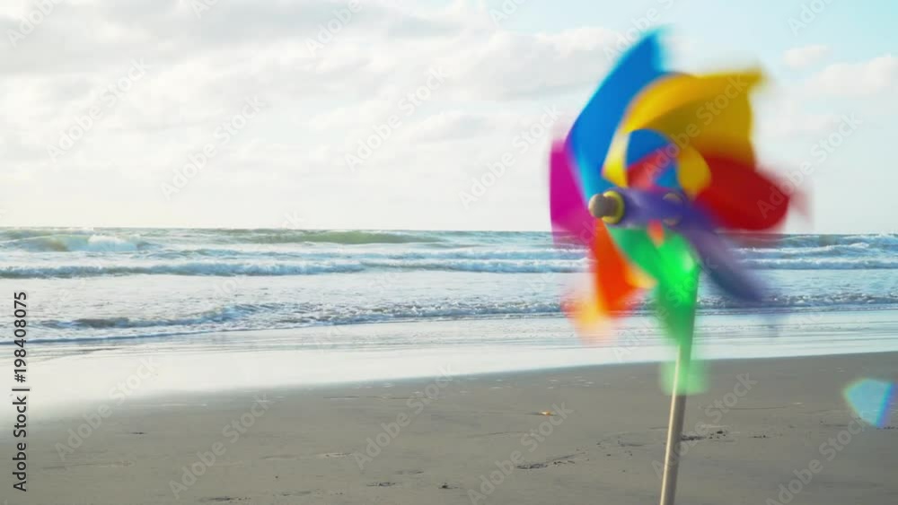 4k. Pin Wheel Toy on the Sandy Beach. Beautiful Ocean, Sunrise. Colorful Background for Greeting Card. Space for Text on Left. Windmill on Blue Sky with White Clouds. Concept Outdoor, Happiness. 4k. Pin Wheel Toy on the Sandy Beach. Beautiful Ocean, Sunrise. Colorful Background for Greeting Card. Space for Text on Left. Windmill on Blue Sky with White Clouds. Concept Outdoor, Happiness.