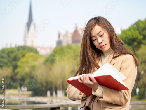 Portrait of beautiful Chinese girl standing and reading a book in wind with blur Gothic architecture background. Happy reader in college.