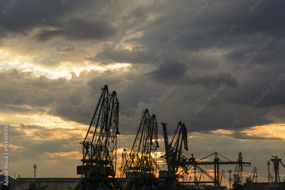 Fototapeta premium Silhouette of port cranes against a dramatic sky at sunset.