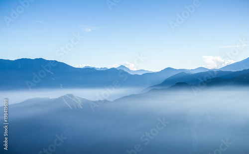 Hazy blue mountains of Zhushan inside Alishan Recreation Area in Taiwan covered by fog during sunrise in morning with bright winter sky.