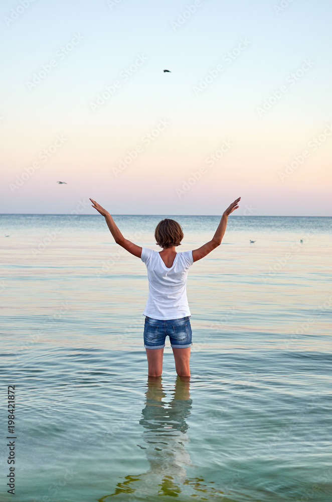 Beautiful young woman standing in water, raising her hands up and flying as seagull on the sea. Girl enjoying freedom and fresh air on ocean beach, copy space