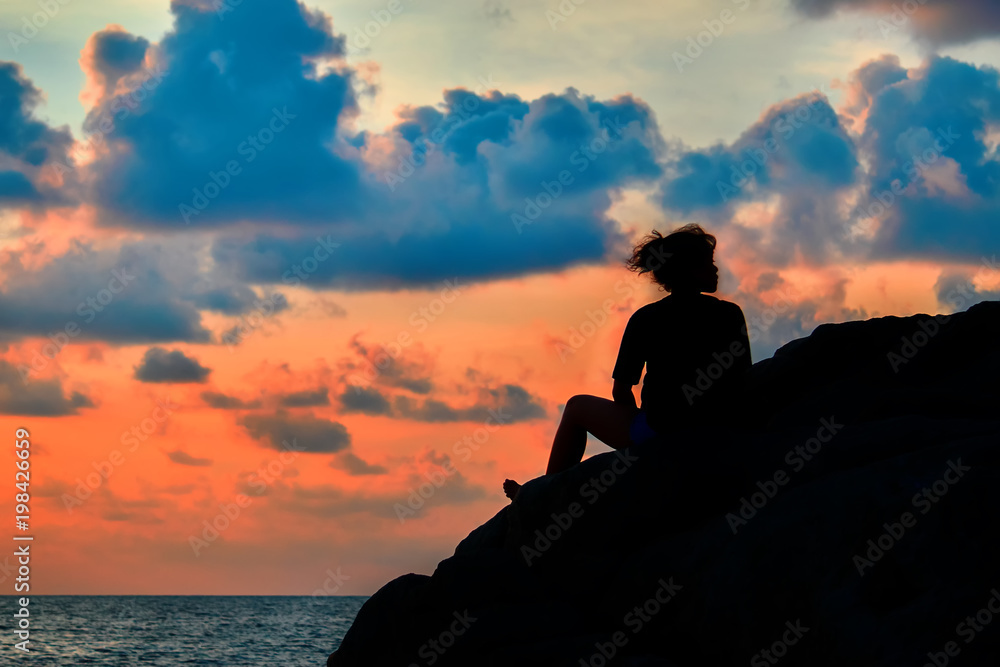Woman silhouette at sunset. Girl sits on  rock above tropical sea. Evening sky of orange color with blue clouds. Landscape.