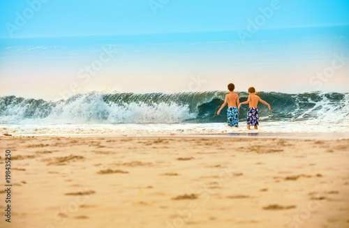 TWO YOUNG BOYS WALKING ON SAND BY LARGE WAVE SURF