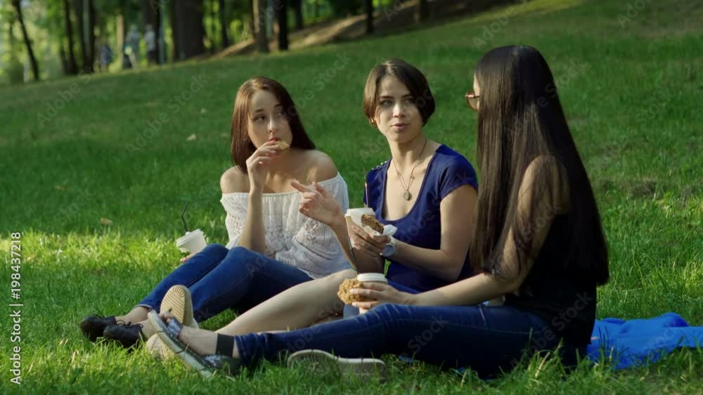 Three young women drinking coffee and eating pastries, sitting on green grass in a summer park. 4K Three young women drinking coffee and eating pastries, sitting on green grass in a summer park. 4K