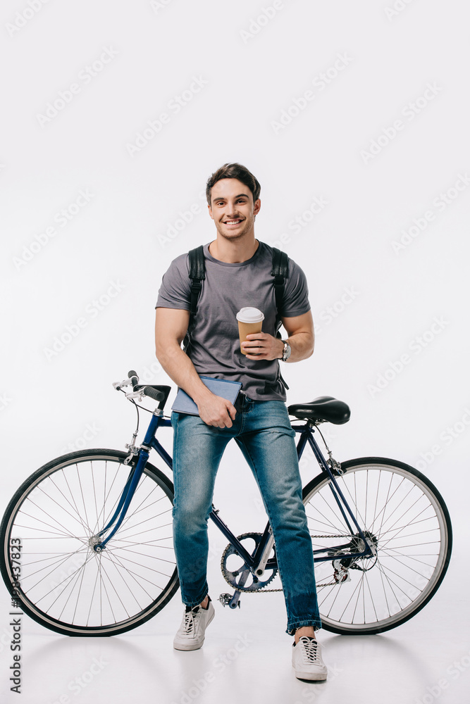 Naklejka premium smiling student holding coffee to go and leaning on bicycle on white
