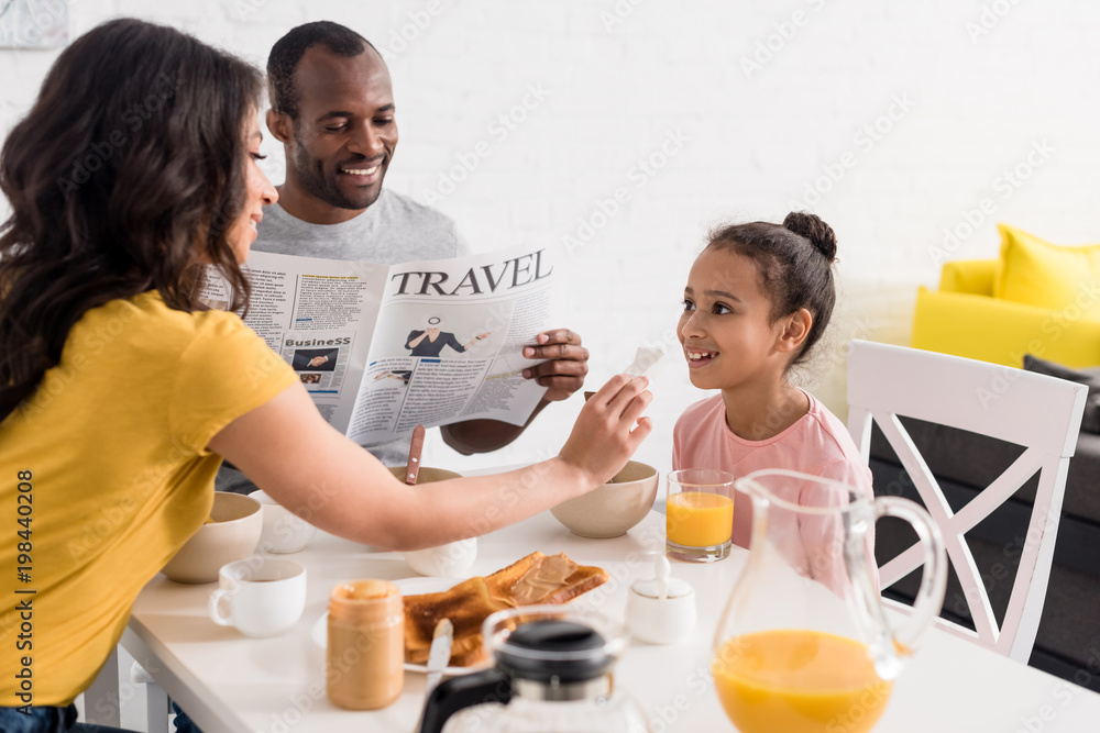 mother wiping face of little daughter after breakfast while father reading newspaper on kitchen