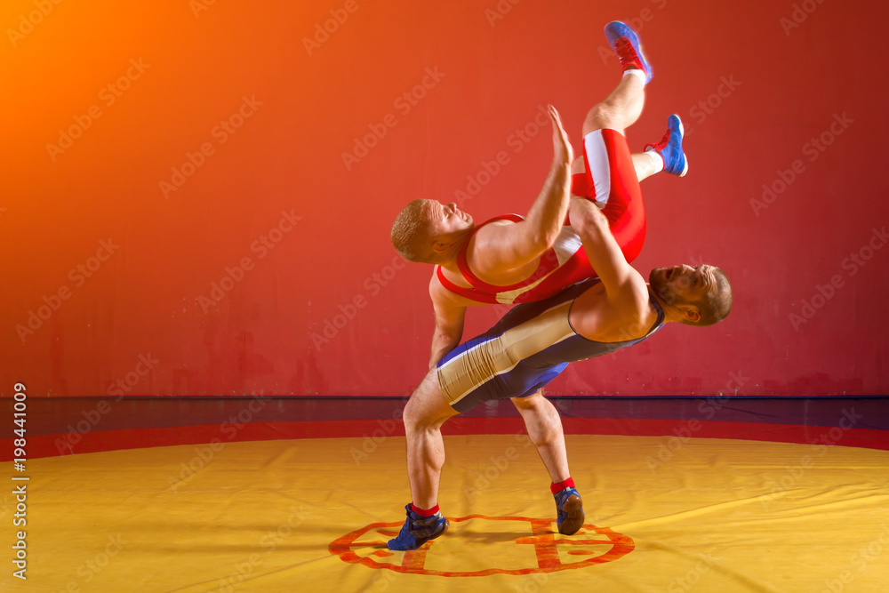 Two young men in blue and red wrestling tights are wrestlng and making a suplex wrestling on a yellow wrestling carpet in the gym Two young men in blue and red wrestling tights are wrestlng and making a suplex wrestling on a yellow wrestling carpet in the gym