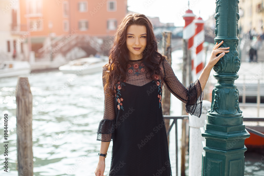 Fototapeta premium Attractive young romantic woman standing on the pier against beautiful view on venetian chanal with boats and gondolas in Venice, Italy. Travel tourist girl on vacation walking happy by Grand Canal